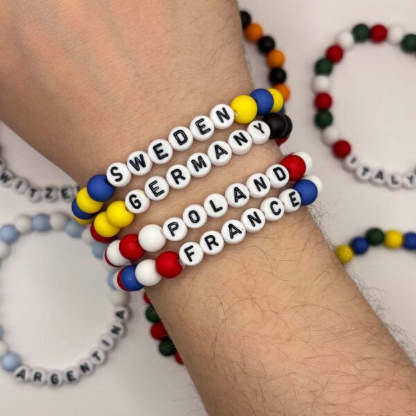 Colorful beaded bracelets on a wrist; white-letter beads spell country names like SWEDEN, GERMANY, FRANCE and POLAND.