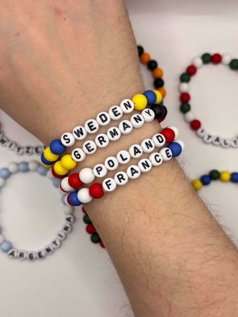 Colorful beaded bracelets on a wrist; white-letter beads spell country names like SWEDEN, GERMANY, FRANCE and POLAND.