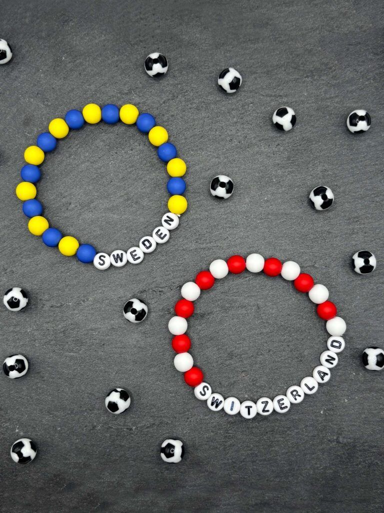 Beaded bracelet spelling SWEDEN in blue and yellow beads, surrounded by small soccer balls on a gray background.