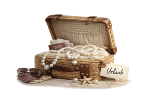 Open wicker suitcase filled with a string of pearls, jewelry, passport, cash, sunglasses, and a sign reading 'Urlaub' on a sandy surface.
