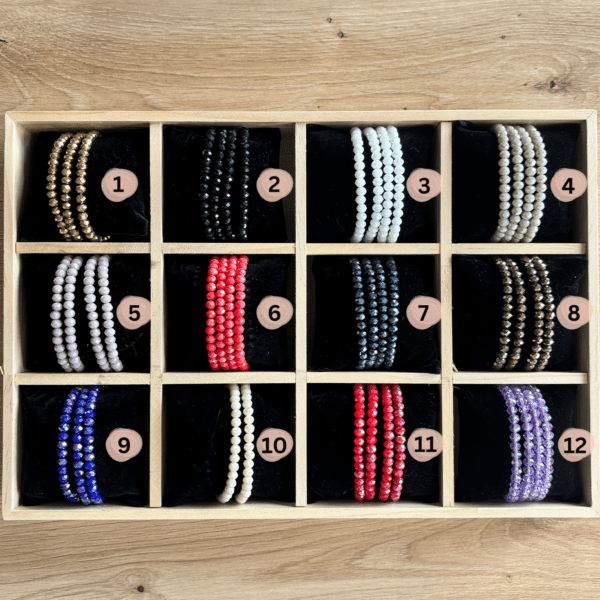 Wooden display tray with twelve compartments holding color-varied beaded bracelets, labeled 1–12 in soft pink circles.