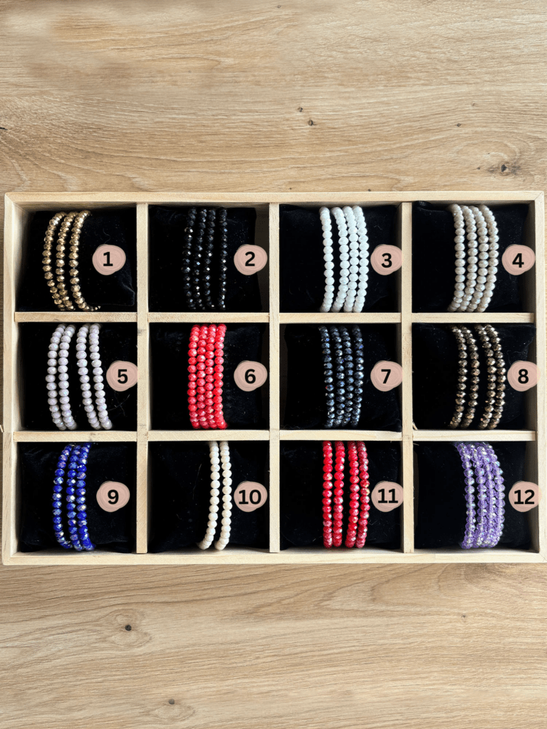 Wooden display tray with twelve compartments holding color-varied beaded bracelets, labeled 1–12 in soft pink circles.