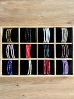 Row of beaded bracelets arranged in a wooden display box with black velvet compartments, showing gold, black, white, red, blue, and purple strands photographed from above on a wooden surface.