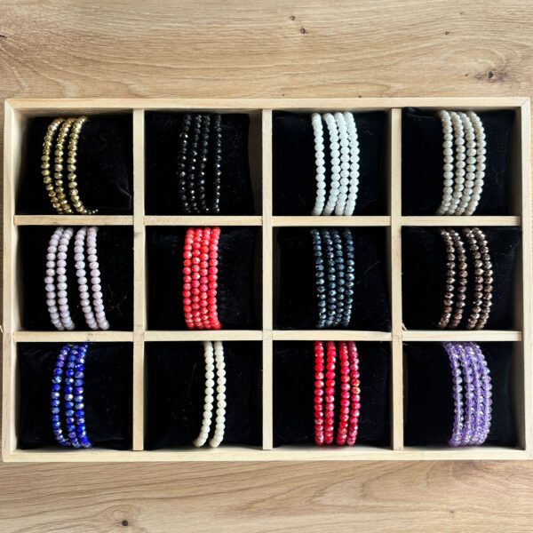 Row of beaded bracelets arranged in a wooden display box with black velvet compartments, showing gold, black, white, red, blue, and purple strands photographed from above on a wooden surface.