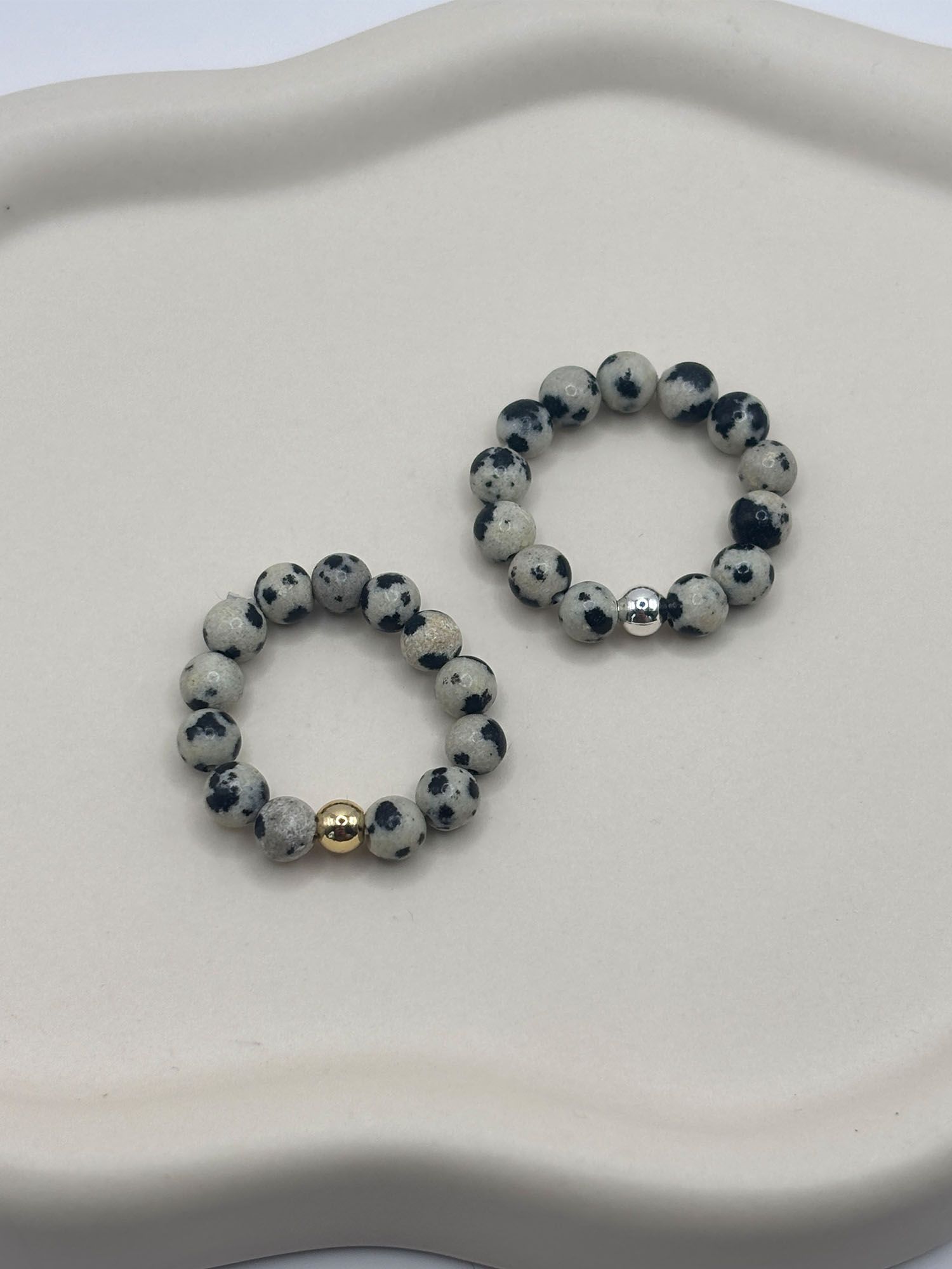 Perlenring_Dalmatiner_Jaspis_Vollansicht_Liegend_Teller Beaded bracelet with speckled gray and black stones and a gold accent bead on a white surface.