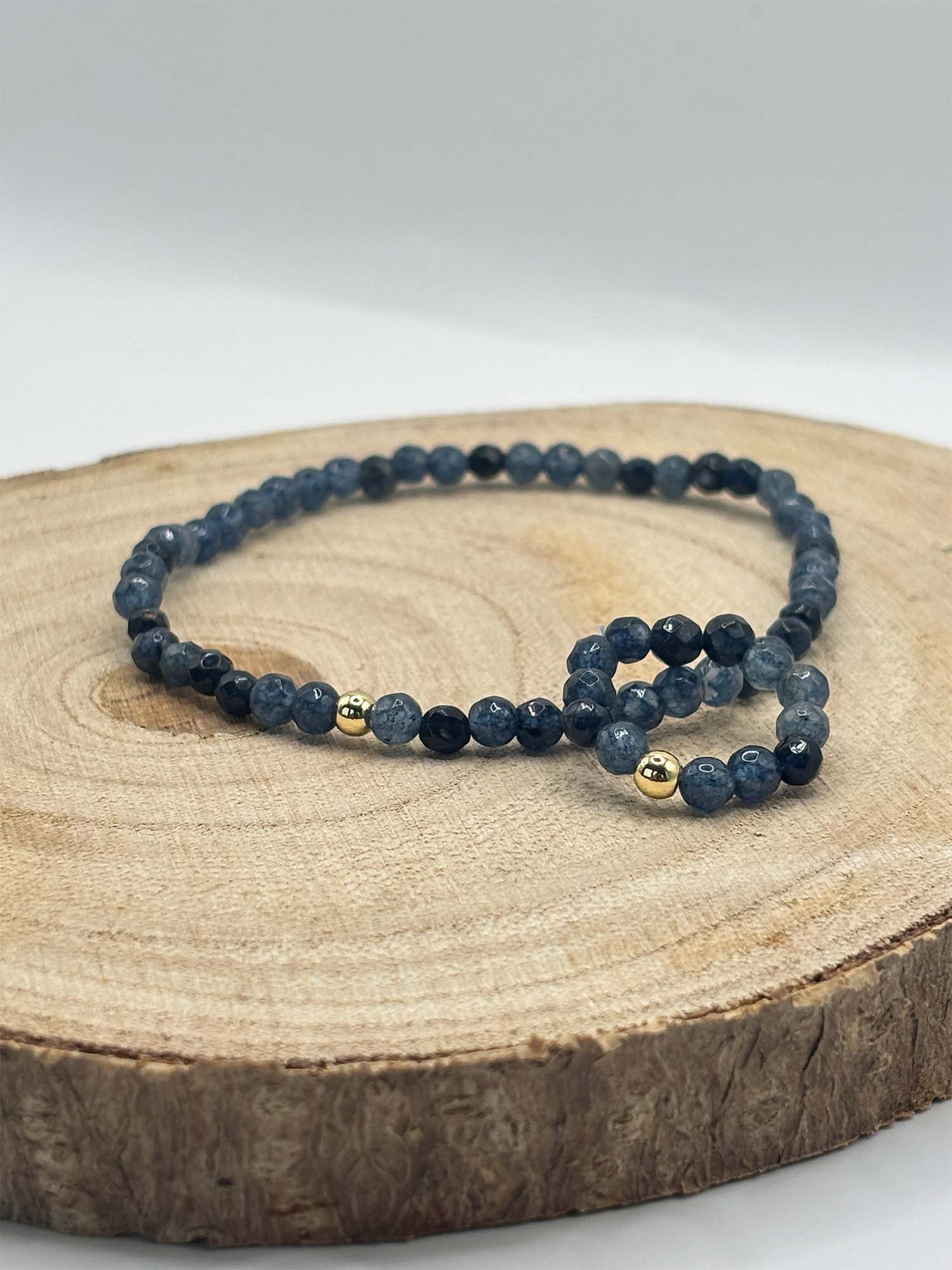 Set_Perlenarmband_Ring_Saphirblau_Jade_mit_Goldperle_Holzplatte_Vollansicht Blue beaded bracelet with gold spacers resting on a wooden tree slice, close-up product shot